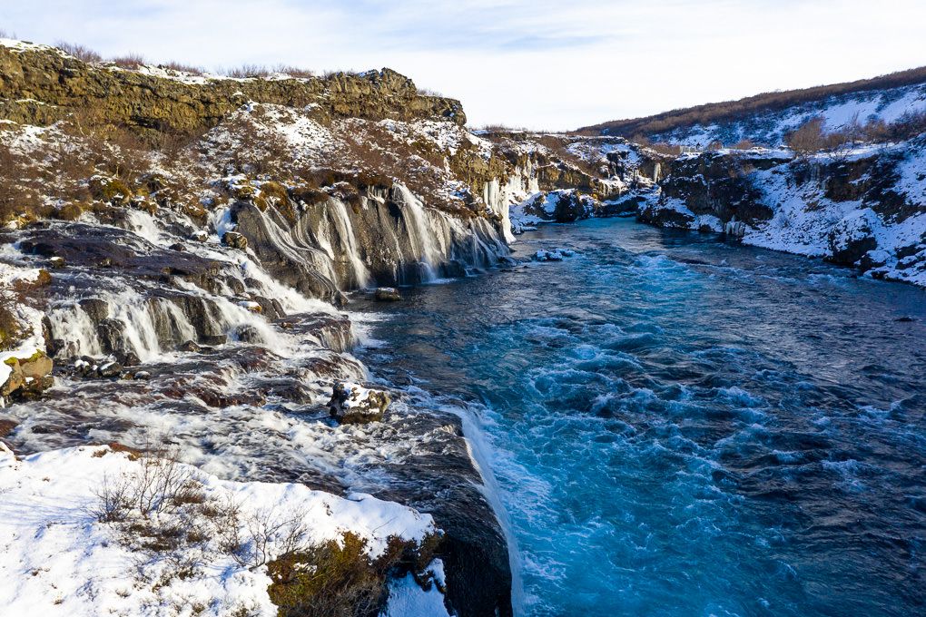 Snow-covered rocky cliffs surround the Hraunfossar Waterfalls in Iceland.
