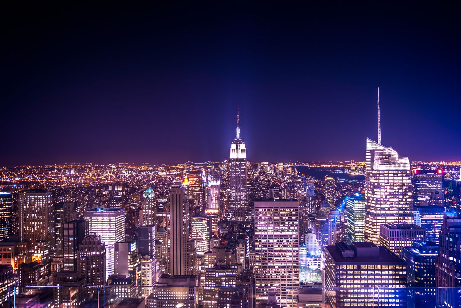 Illuminated Empire State Building and skyscrapers in Manhattan skyline against dark blue night sky with city lights.