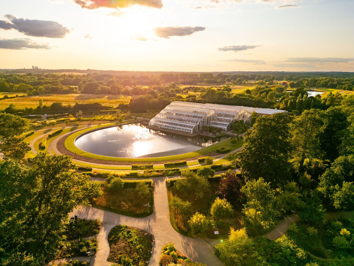 Aerial drone photograph of a modern glasshouse and ornamental lake surrounded by landscaped gardens and rolling countryside at sunset.