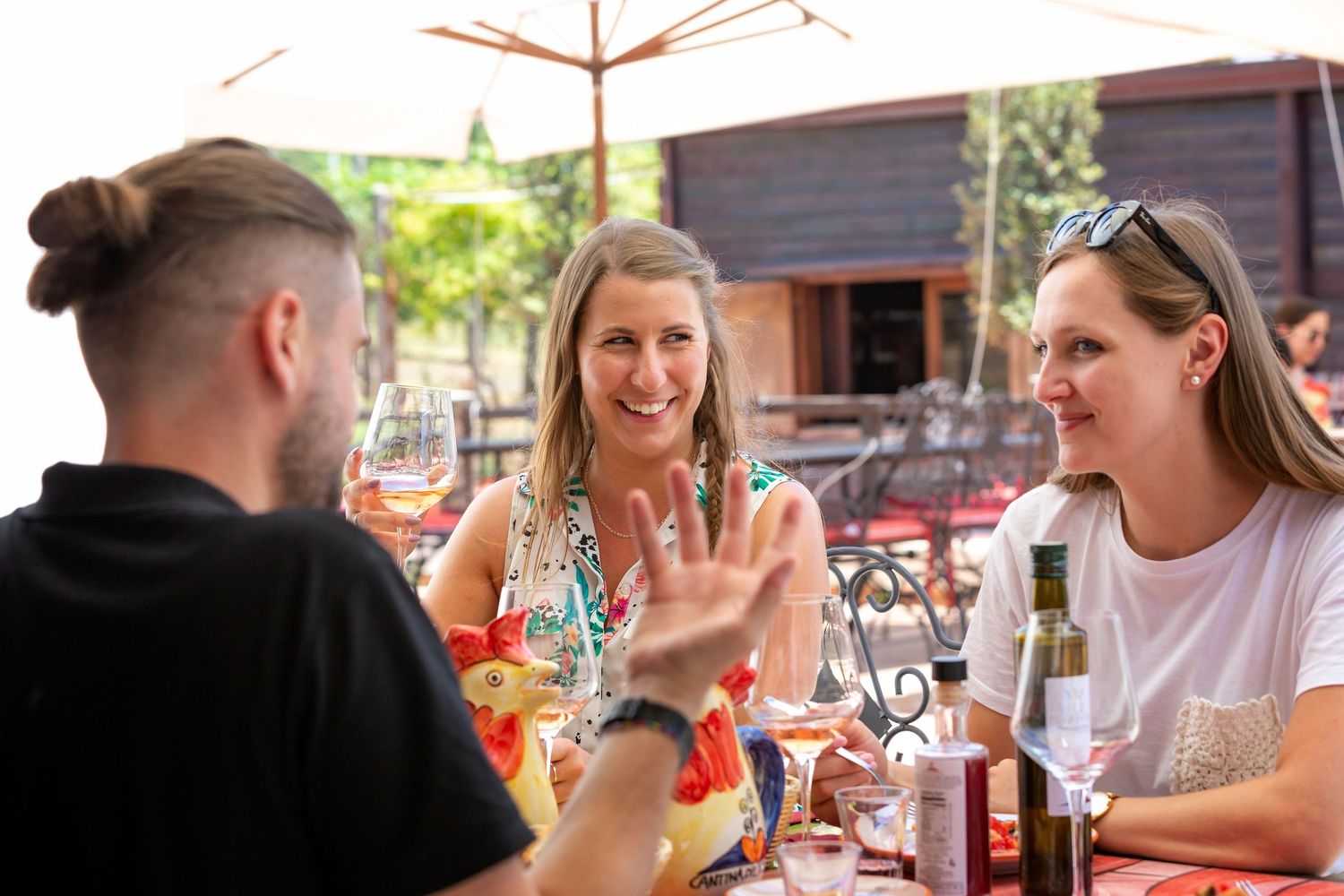 Three friends laughing and chatting at an outdoor restaurant table with drinks and a bottle of wine under a patio umbrella.