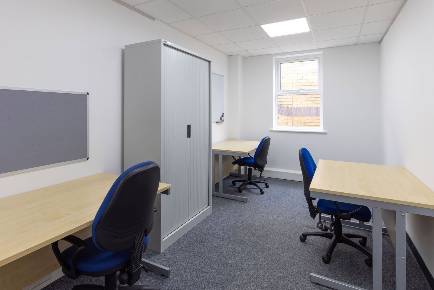 Small office space with three blue chairs, light wood desks, gray storage cabinet, whiteboard, and carpeted flooring.