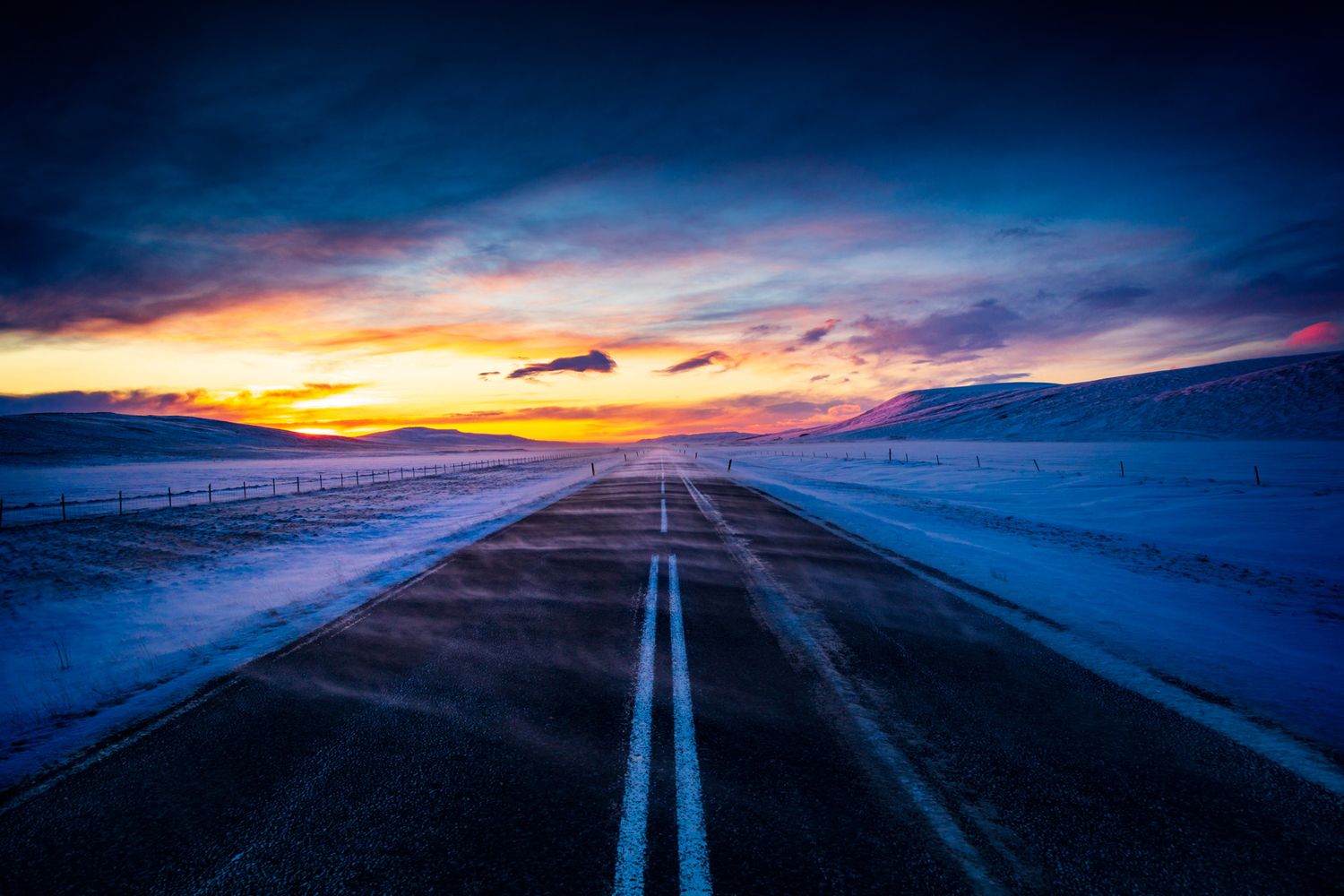 Dramatic sunset illuminates a snowy two-lane highway stretching into the horizon under colorful winter skies.