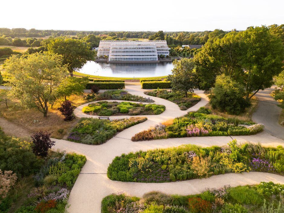 Aerial view of winding garden paths leading to a large greenhouse surrounded by lush landscaping and colorful flower beds.