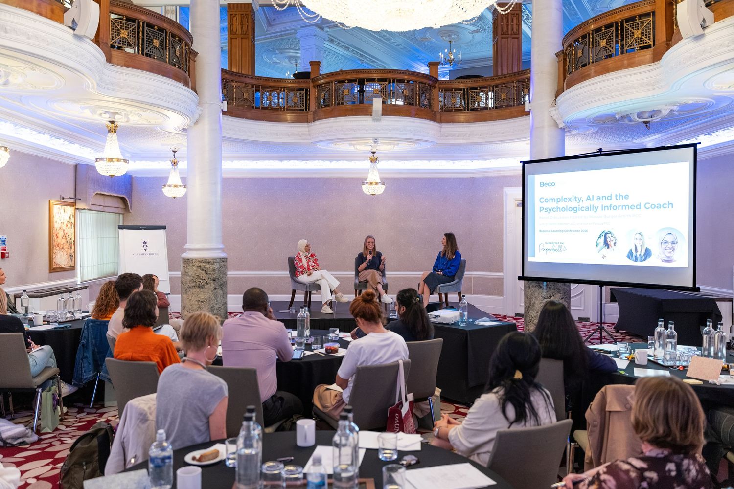 Panel discussion at an elegant conference venue with audience seated at round tables facing three speakers on a stage.