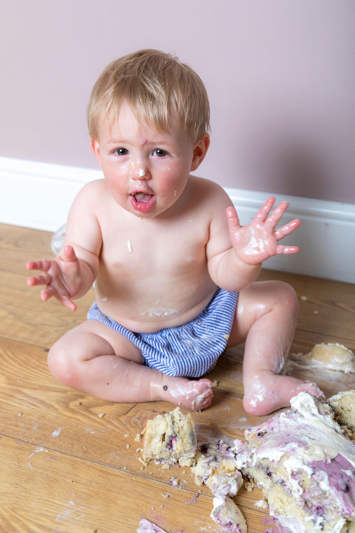 Messy toddler boy sitting on floor covered in birthday cake smash, hands raised with frosting.