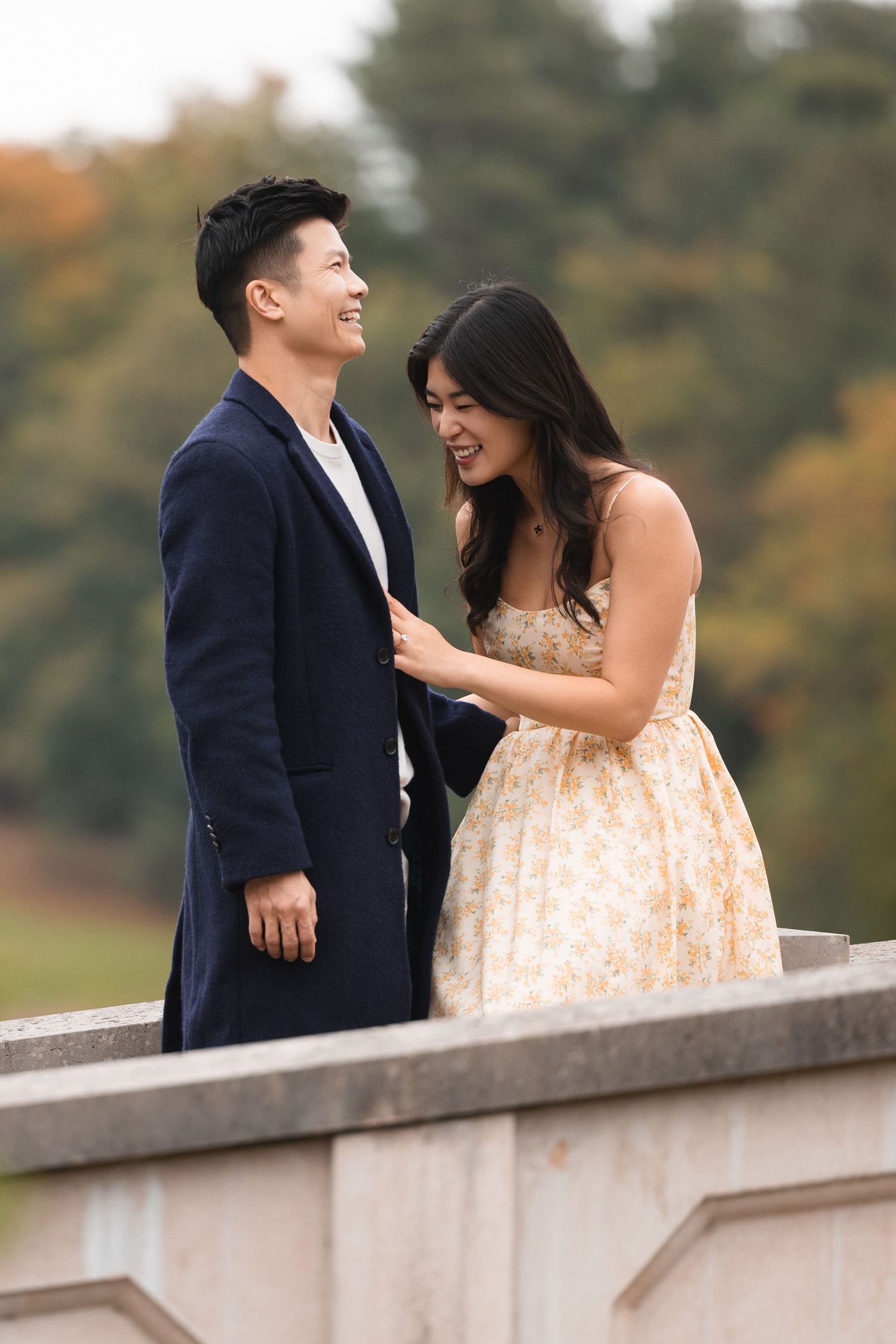 A smiling couple in elegant attire shares a joyful moment outdoors, with autumn trees in the background.