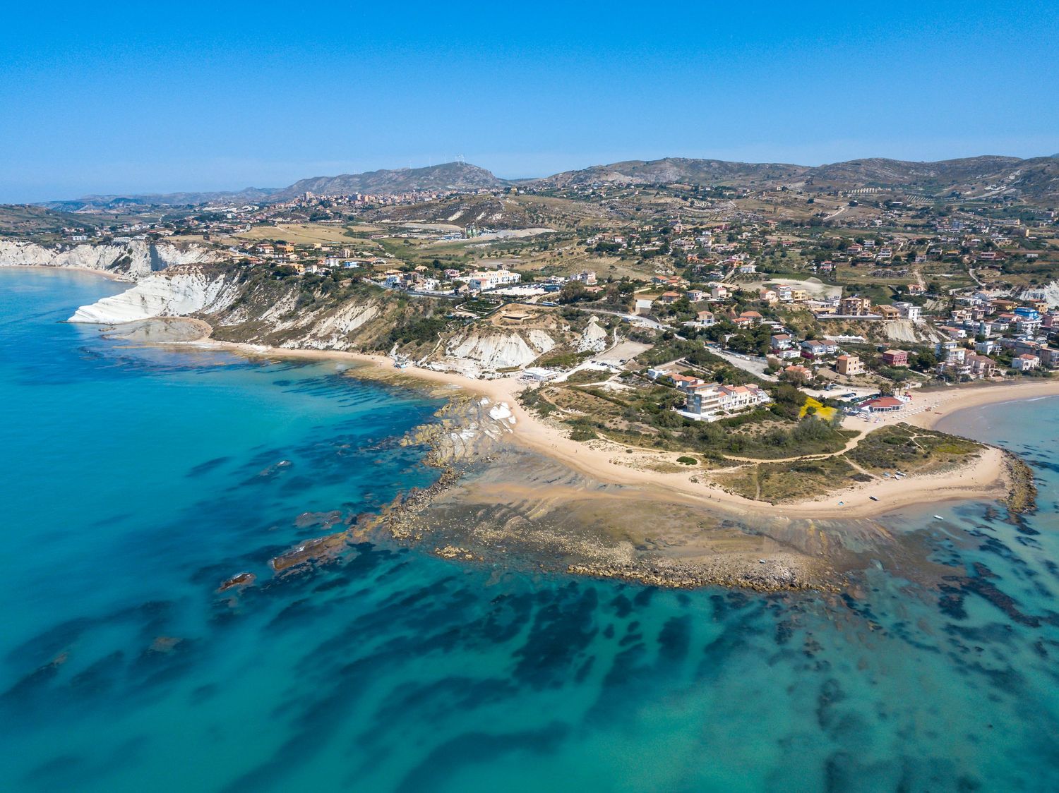Aerial view of a stunning coastal village with turquoise waters, sandy beach, white cliffs, and hillside homes under a clear blue sky.