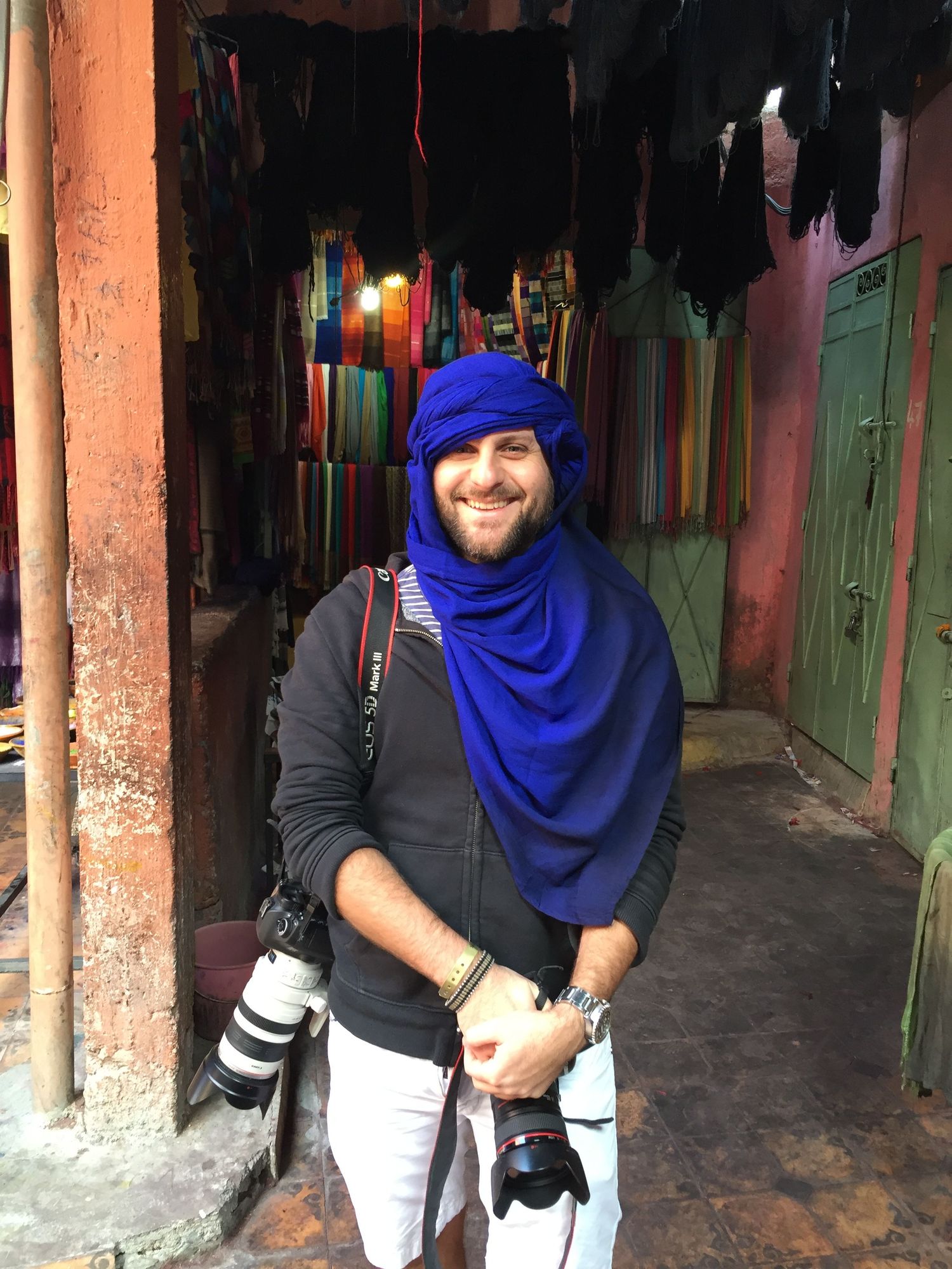 Man wearing blue turban and scarf with camera gear in colorful Moroccan market souk.