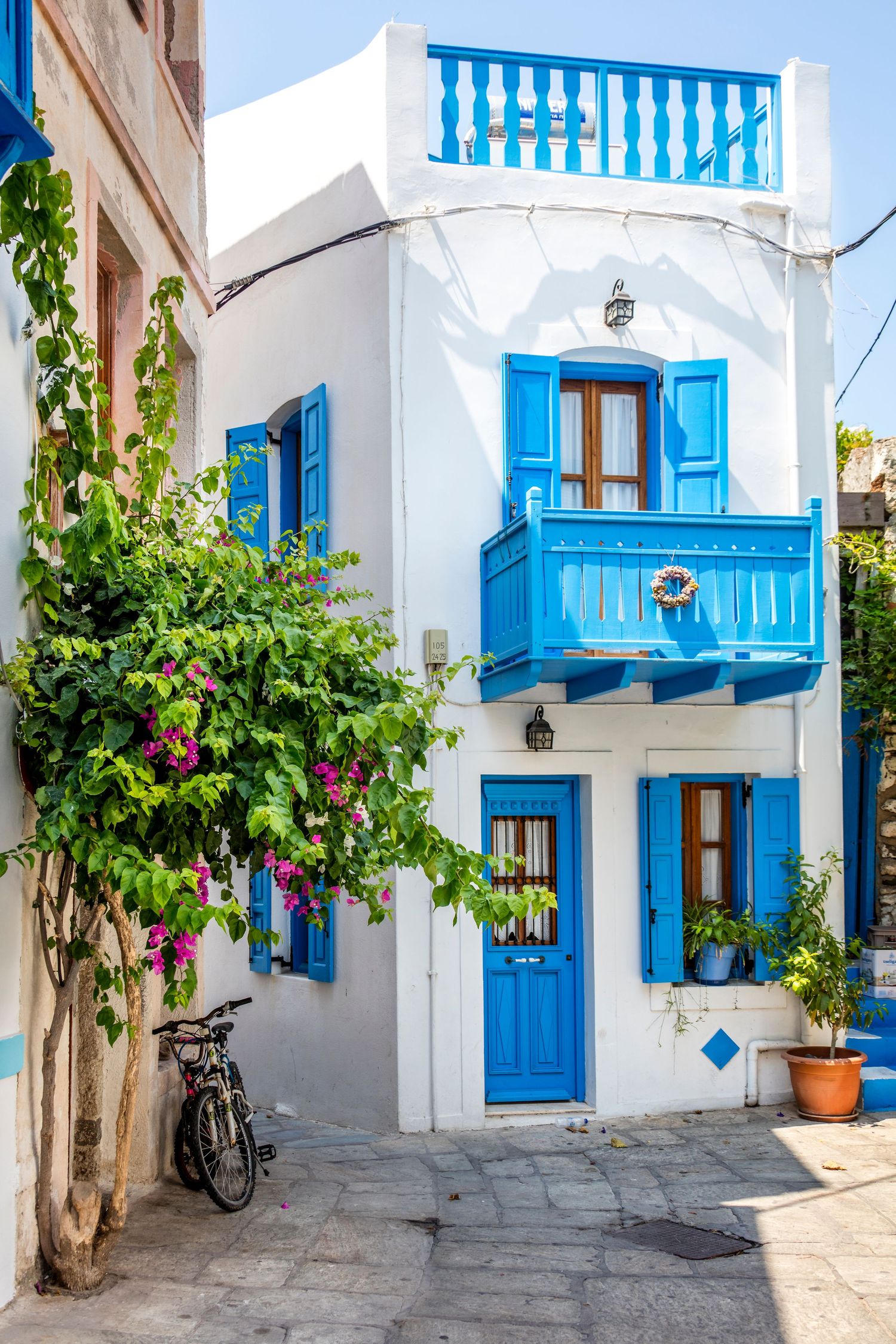 Charming white Greek island building with blue shutters, balcony, and colorful bougainvillea flowers.