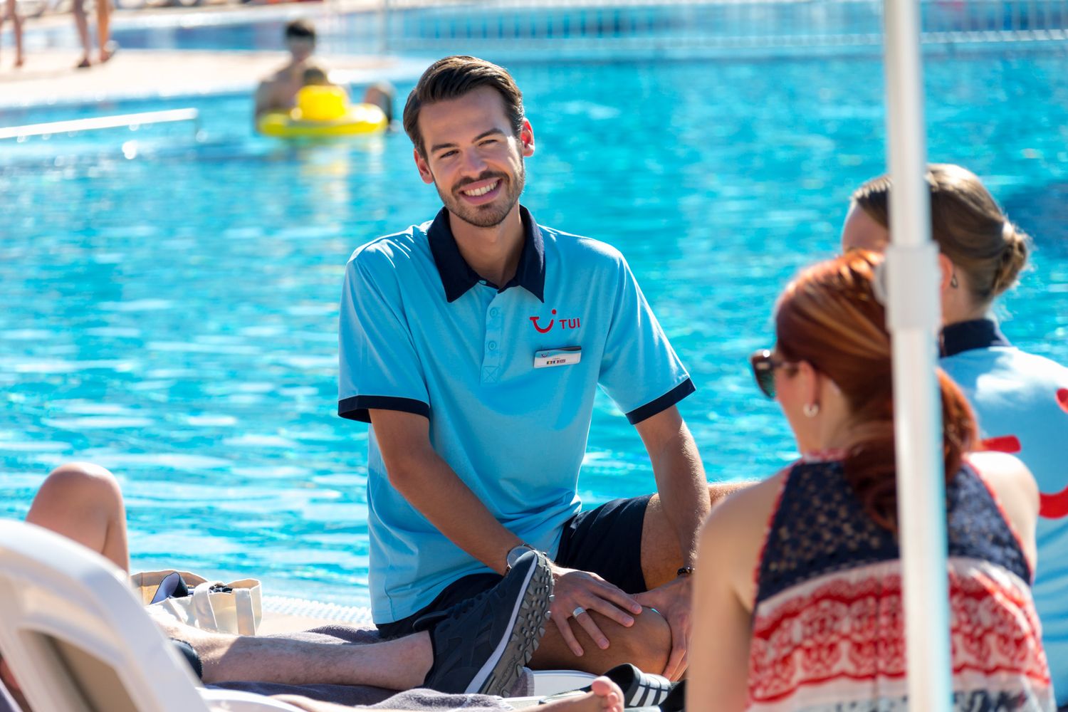 A TUI Staff member in blue polo shirt sits poolside with guests at resort swimming pool during sunny day.