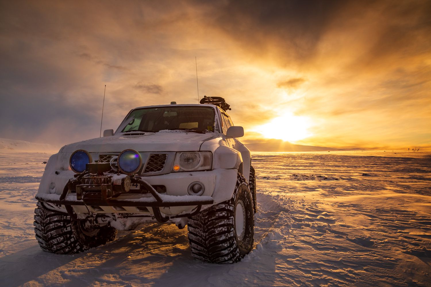 Modified white SUV with large tires parked on a snowy terrain during an epic golden sunset.