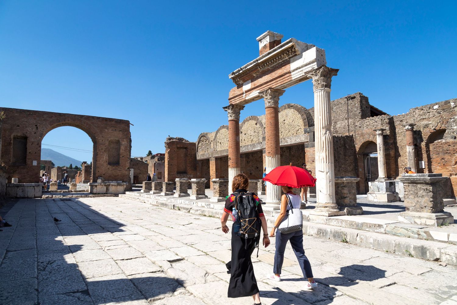 Two tourists, one holding a red umbrella, walk across the ancient stone pavement of the Pompeii Forum past towering Roman columns and ruins.