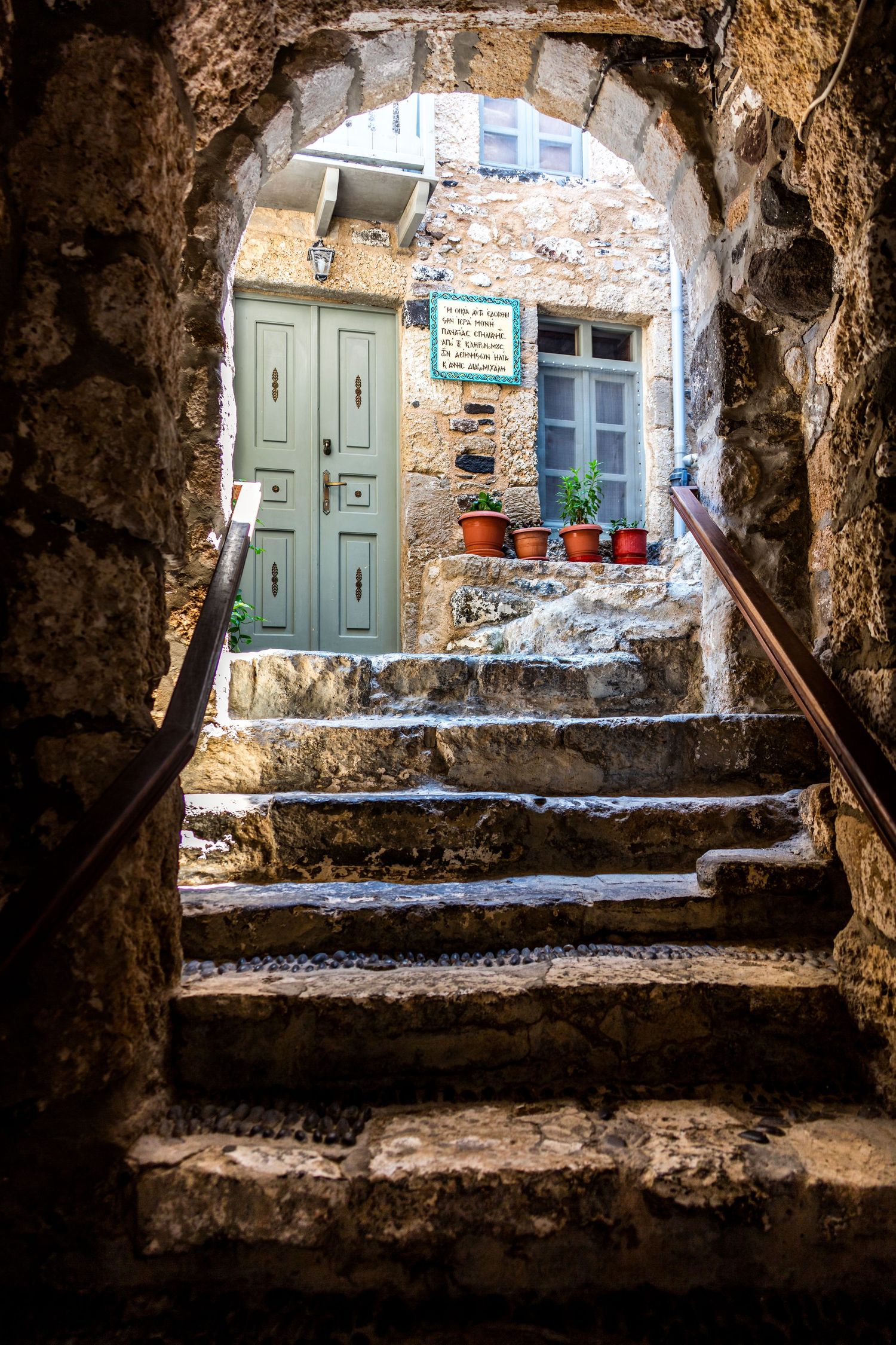 Stone archway framing ancient steps leading to a rustic green door with terracotta pots in a medieval village.