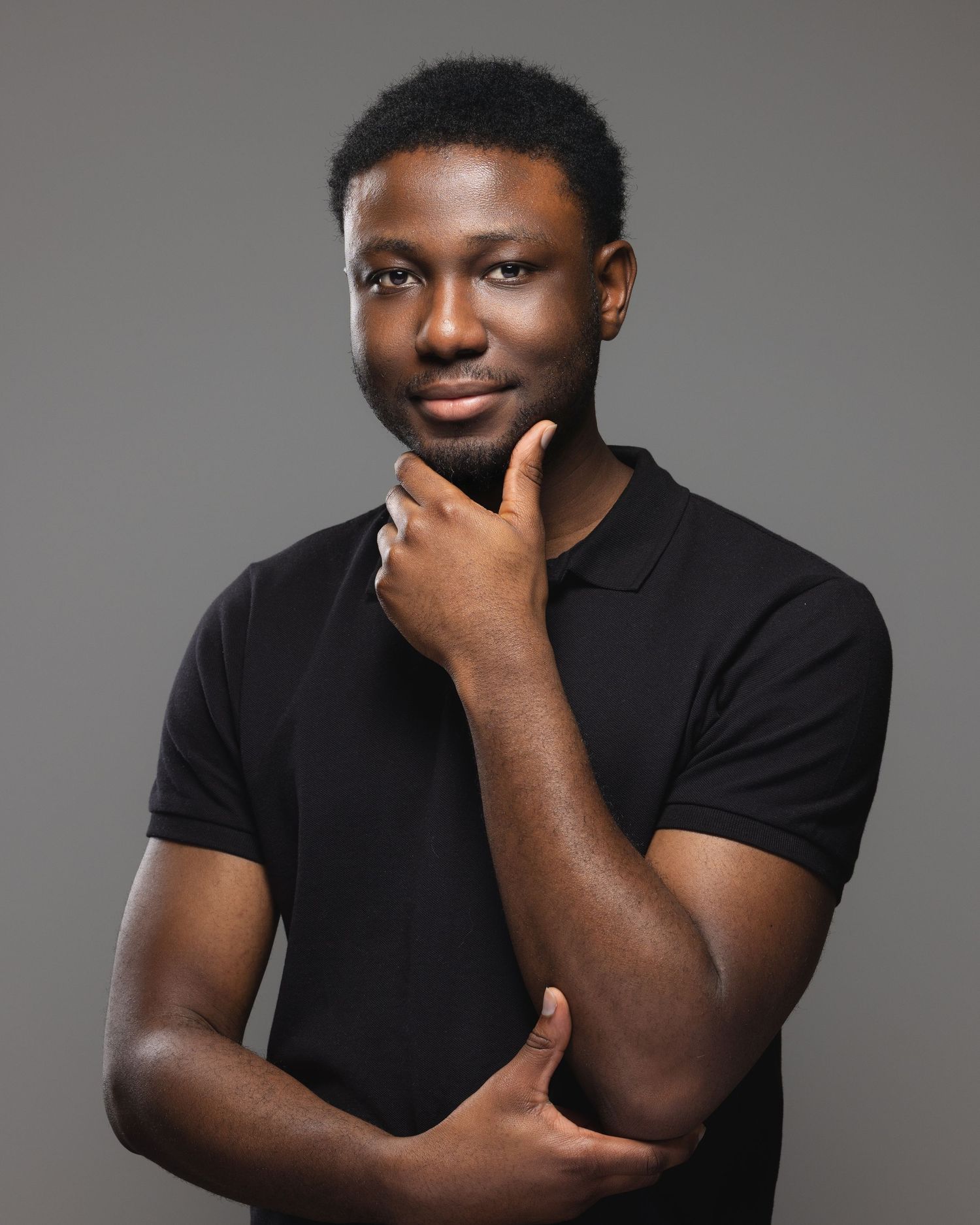 Young Black man in black polo shirt poses thoughtfully with hand on chin against gray studio background.