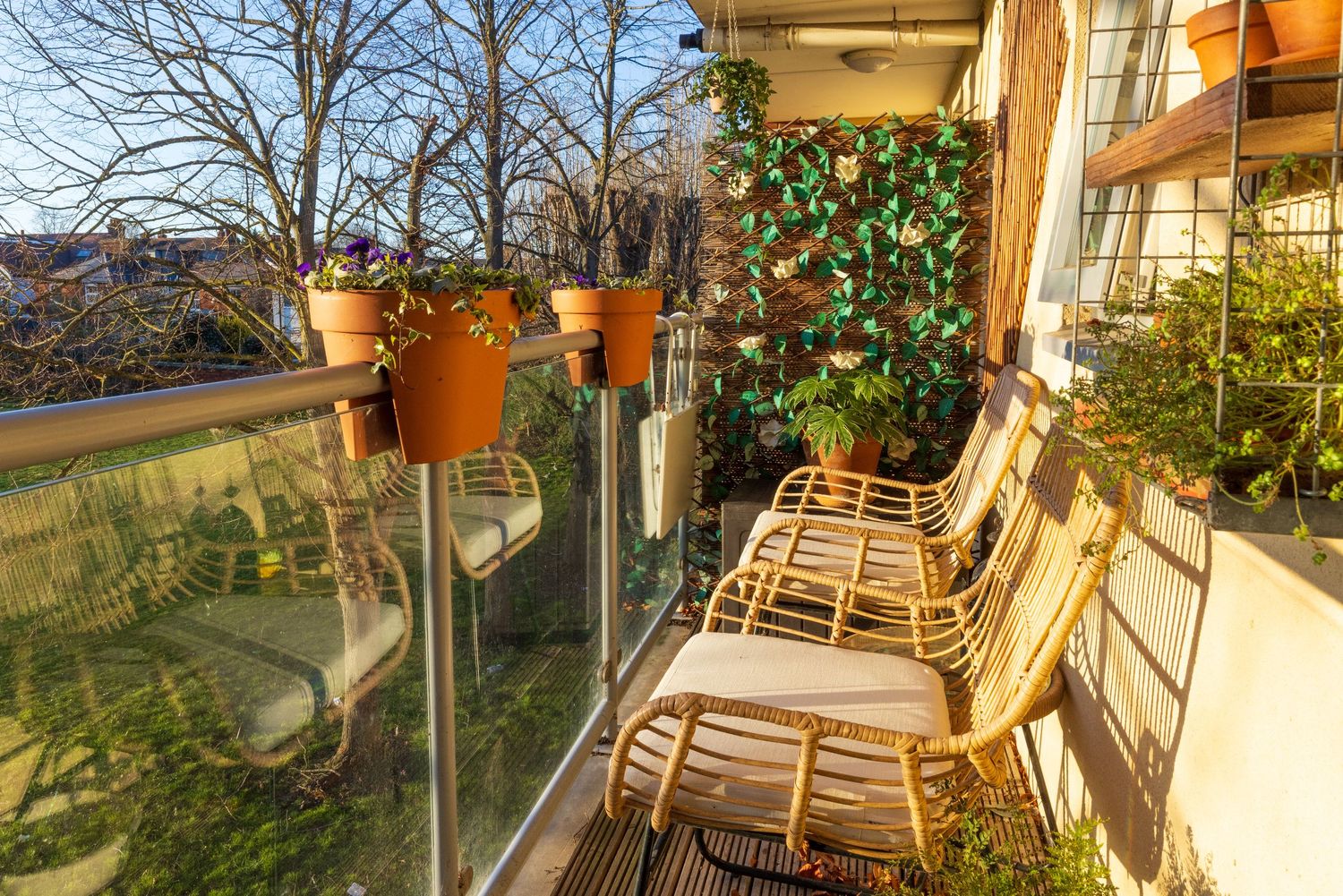 Cozy balcony with rattan chairs, terracotta potted plants, glass railing, and lush green wall decor in warm sunlight.