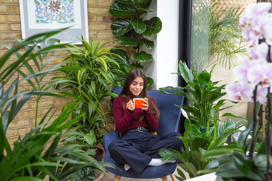 A lifestyle shoot of a girl relaxing in a cosy reading nook surrounded by lush green plants and natural light from a window.
