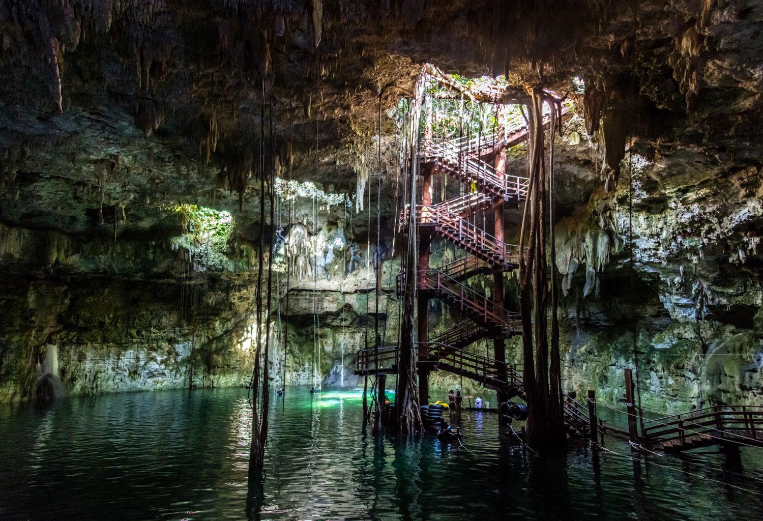 Spiral staircase descends into illuminated cenote cave with turquoise water and limestone formations in Mexico.