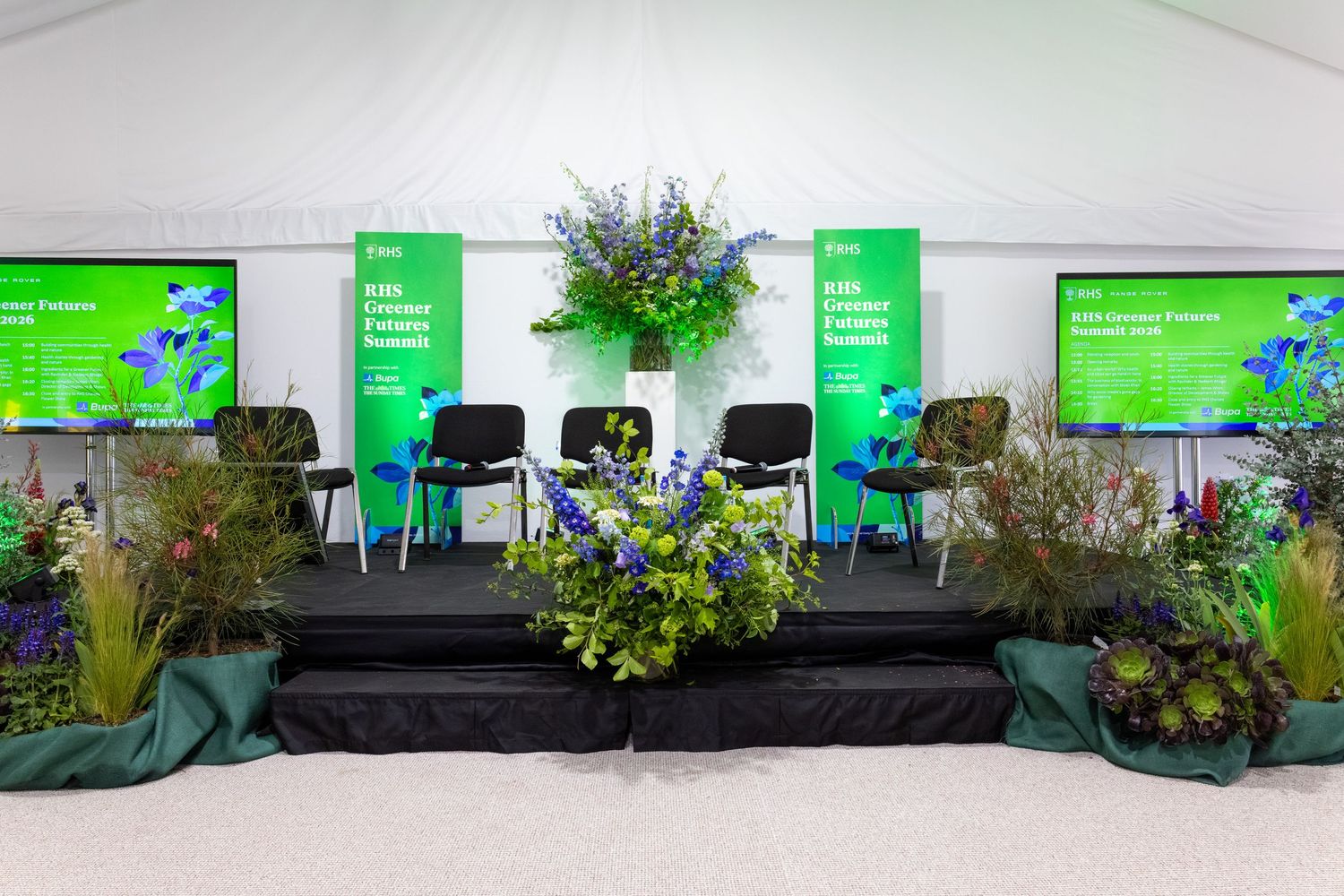 Floral stage display with green banners and colorful flower arrangements at an outdoor event.
