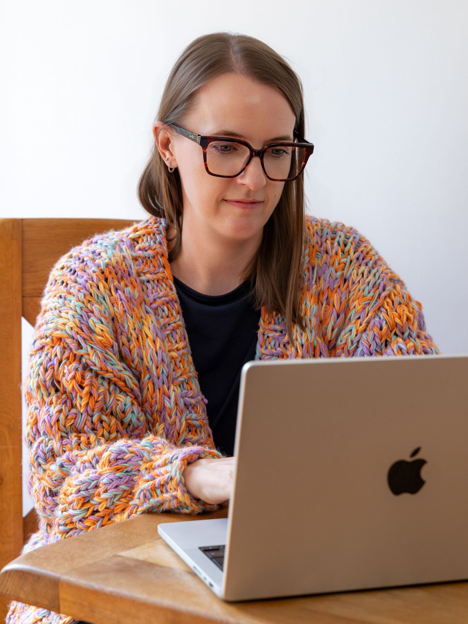 Woman wearing colorful knit cardigan and glasses working on Apple MacBook laptop at wooden desk.