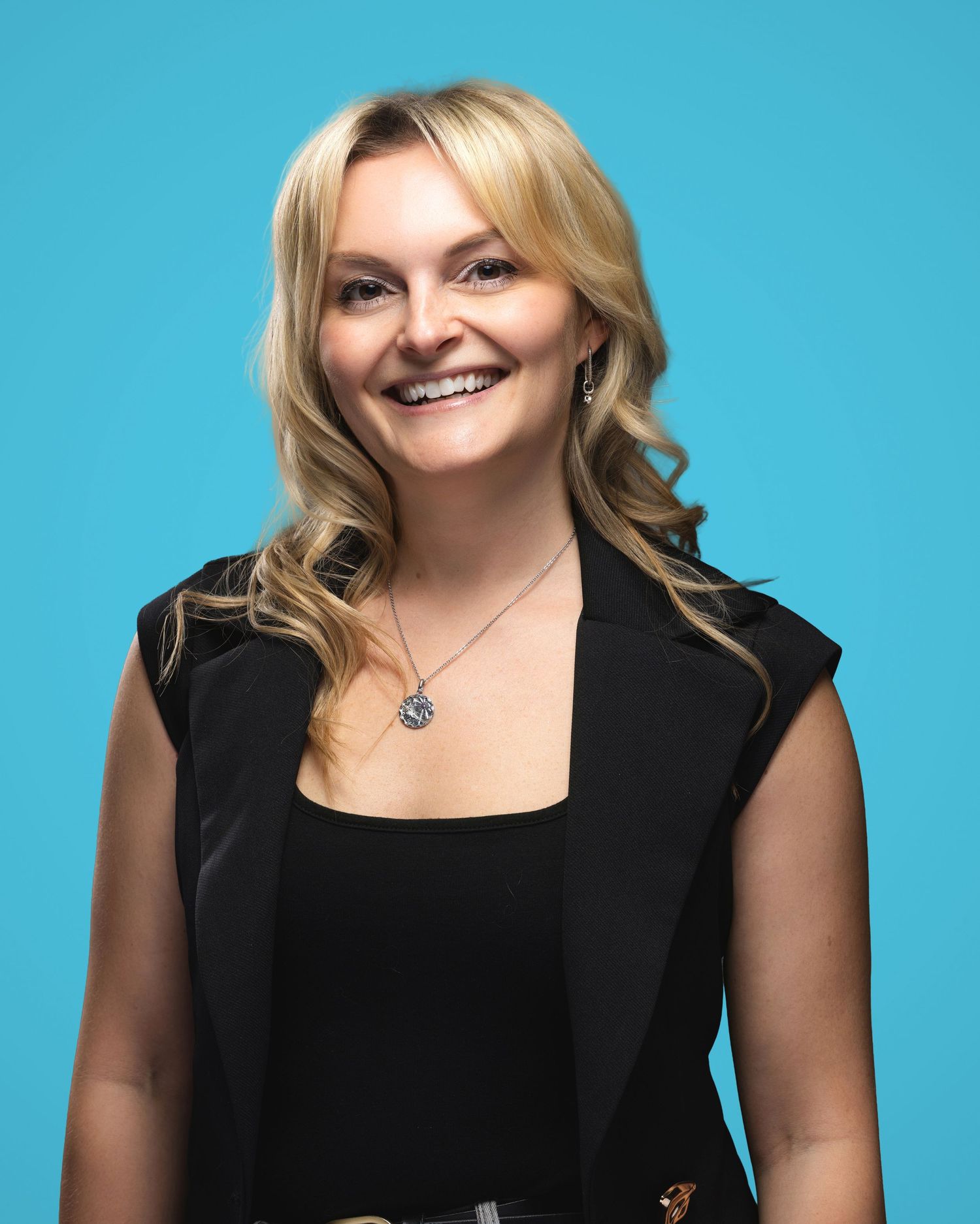 Smiling blonde woman in black sleeveless blazer poses against bright blue background for professional headshot.