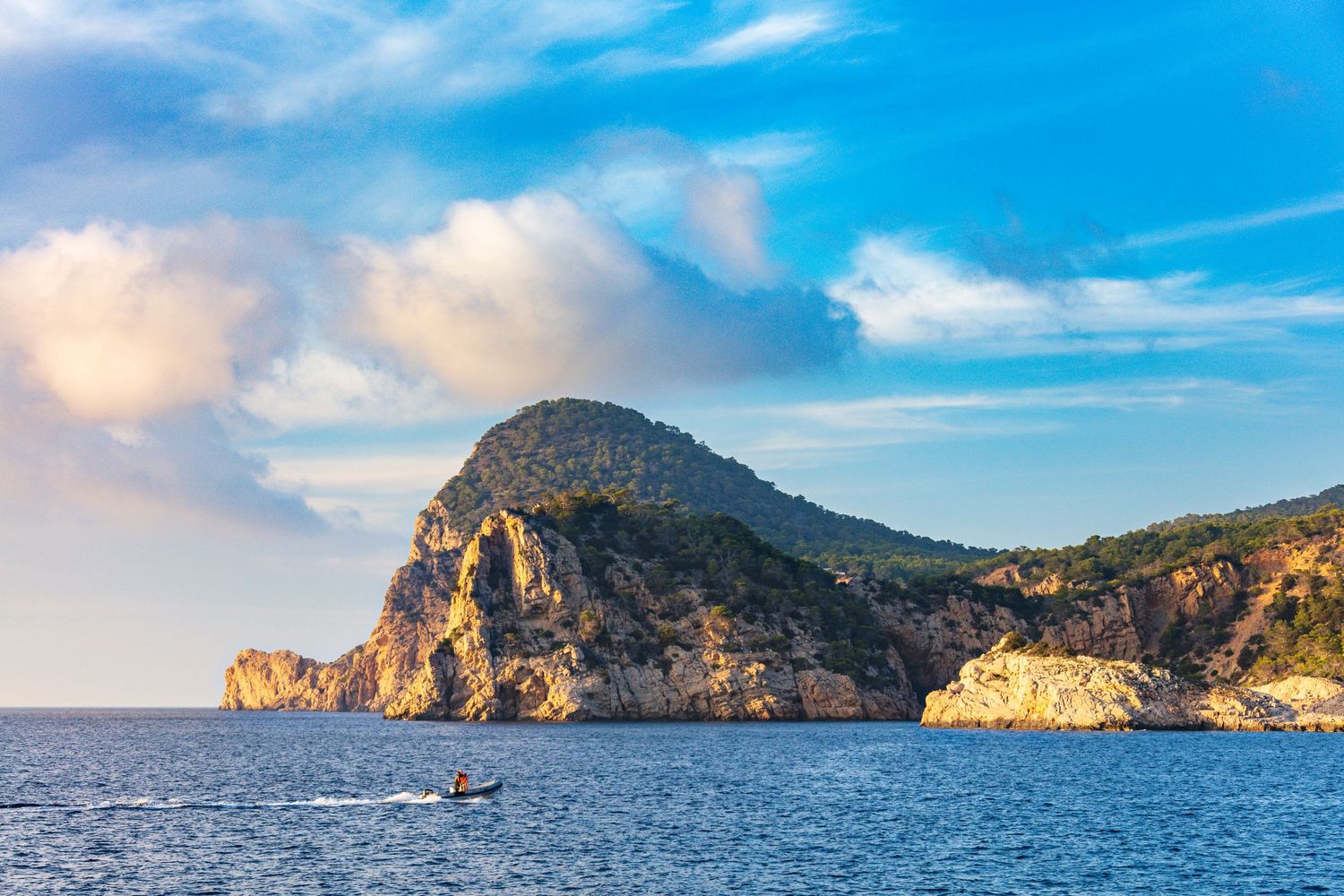Rocky Mediterranean island rising from calm blue sea under dramatic cloudy sky at golden sunset.