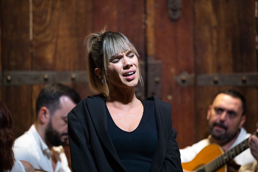 A performer sings passionately on stage while accompanied by musicians with guitars against a wooden backdrop.