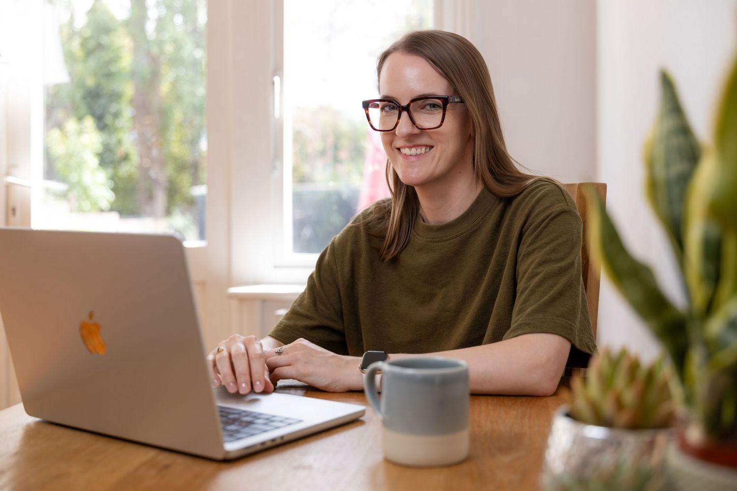 Woman with glasses smiling while working on a MacBook laptop at a wooden desk with a coffee mug and plant nearby.