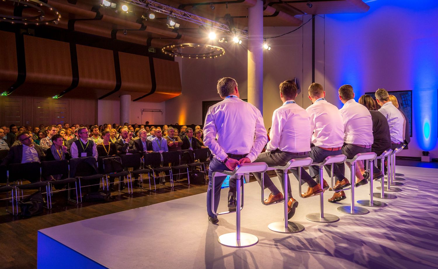 A panel of men in white shirts seated on bar stools face a large audience at a professional conference event.