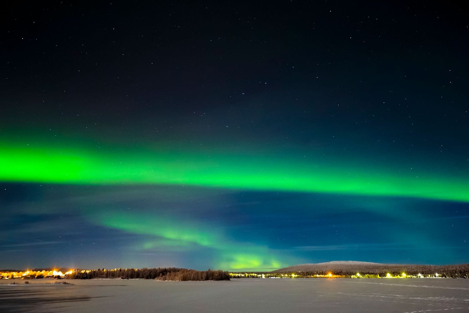 Green aurora above a snowy Arctic landscape with distant town lights.