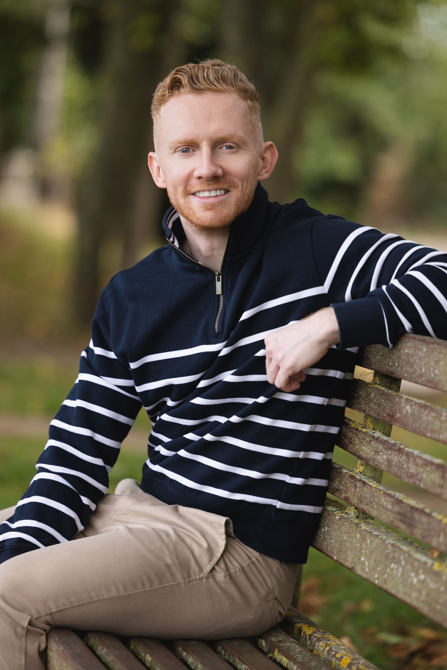 Smiling redhead man in striped navy sweater sitting relaxed on a wooden park bench outdoors.