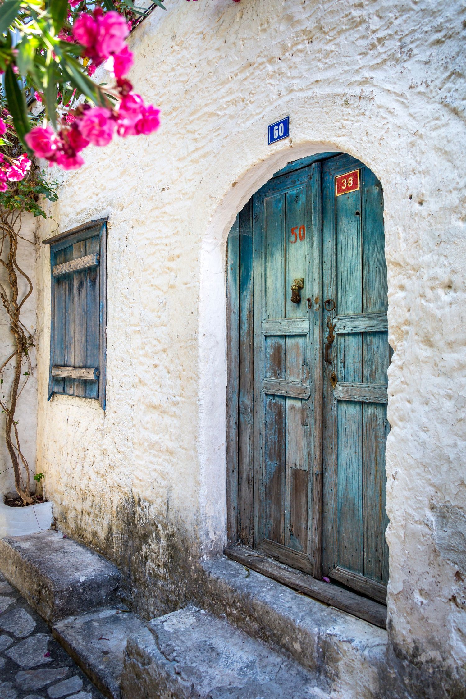 A rustic teal wooden arched door set in a whitewashed stone wall adorned with pink bougainvillea in Greece.