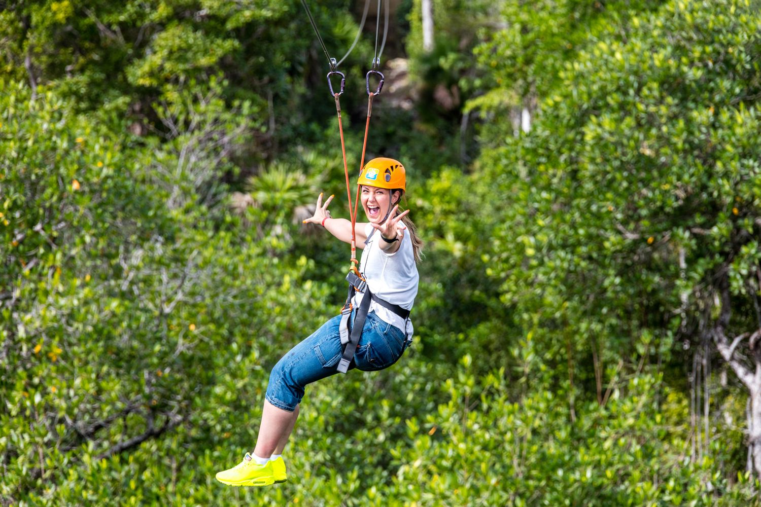 Someone wearing an orange helmet and safety harness ziplining through lush green forest canopy.