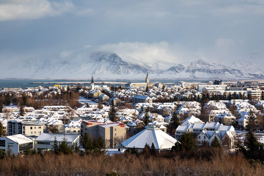 Panoramic winter view of Reykjavik, showing snow-covered rooftops and buildings against a backdrop of mountains.