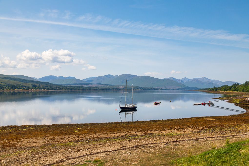 Panoramic view of a Scottish loch near Fort William with mountains in the background and boats along the shoreline under blue skies.
