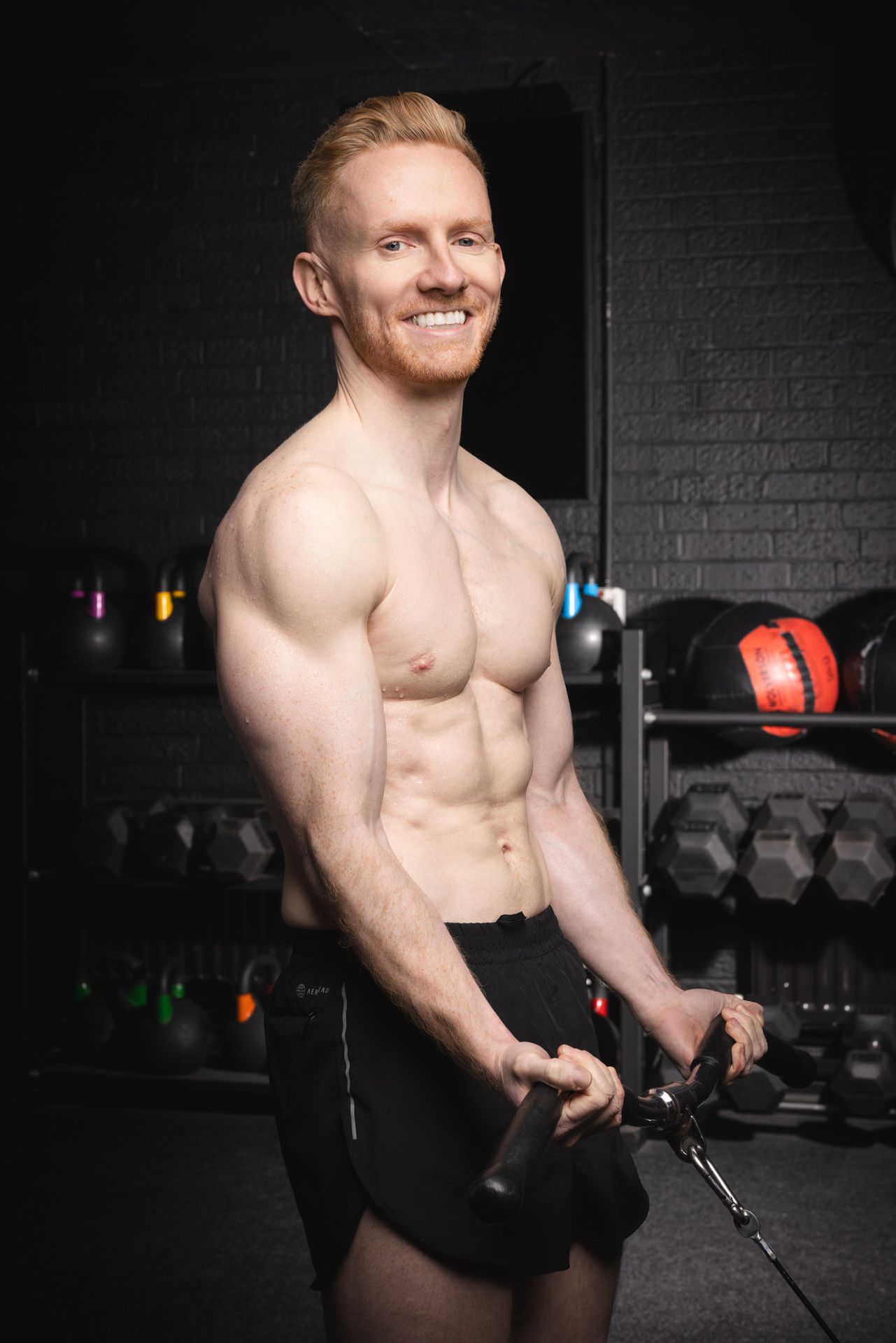 Athletic male with muscular physique posing shirtless in a Hertfordshire gym with fitness equipment in the background.
