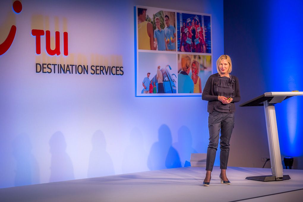 Conference presenter on stage with swooping blue lighting and multiple screens displaying TUI branding in background.