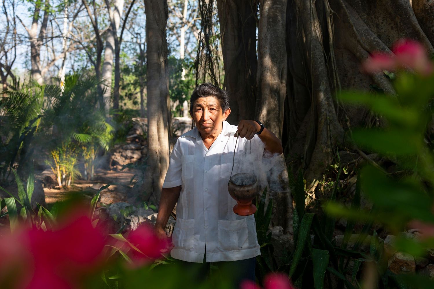 Man in white guayabera shirt holding a gourd in a lush tropical garden with pink flowers and large trees.