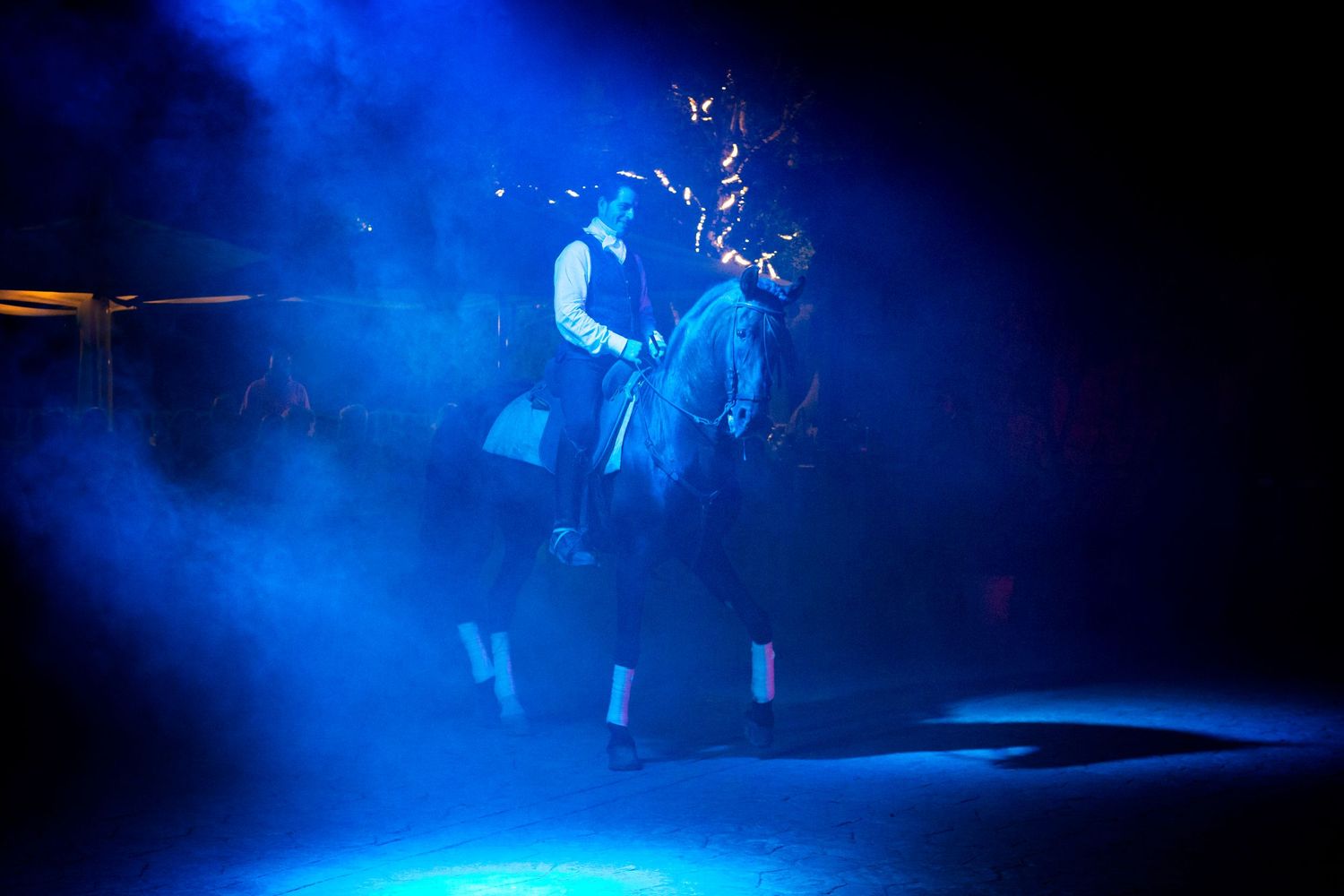 Rider in white shirt and vest performs on a rearing horse in a dark arena, lit by dramatic blue stage lighting and drifting haze.