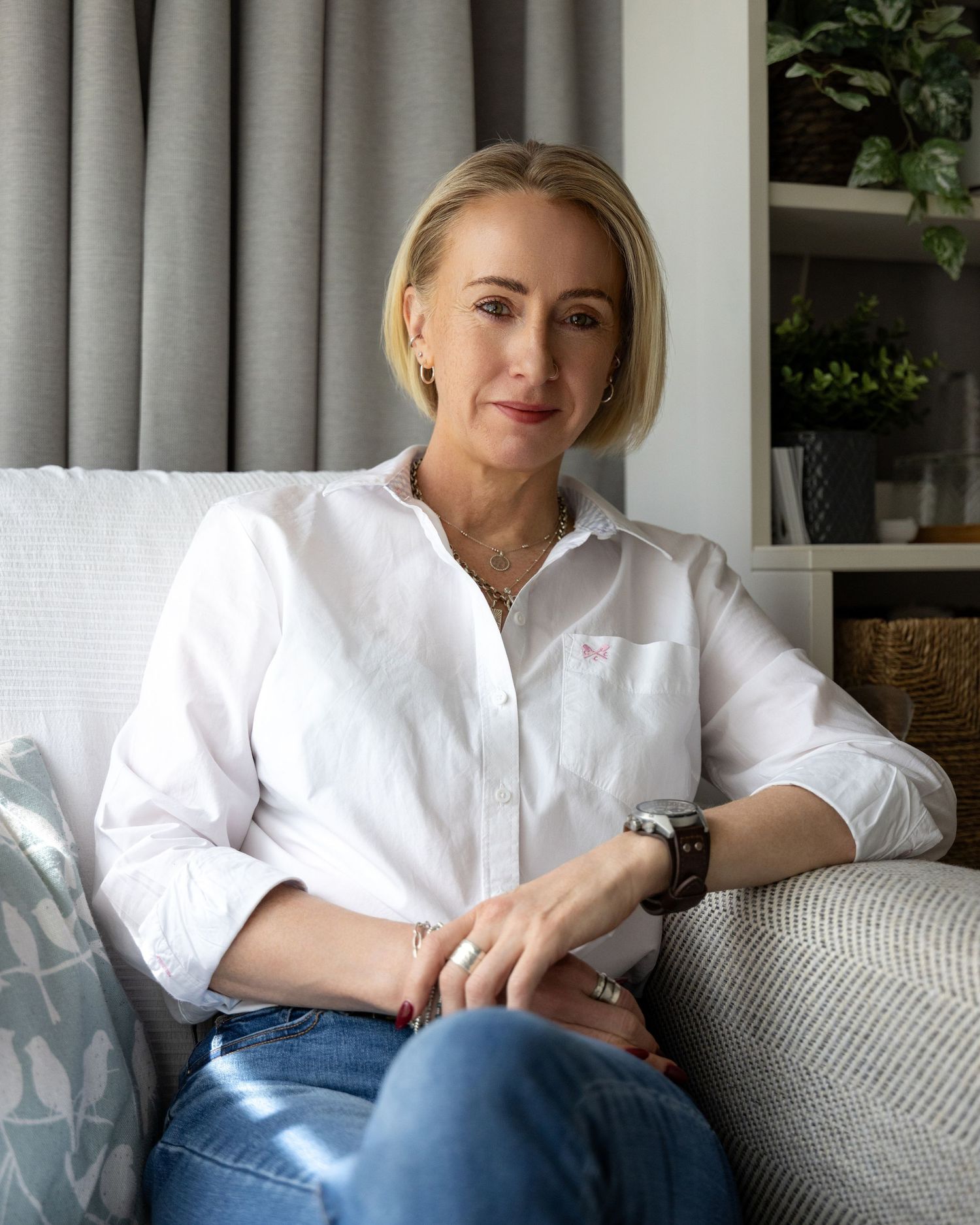Blonde woman in white shirt and jeans sits relaxed on a light gray sofa in a modern living room.