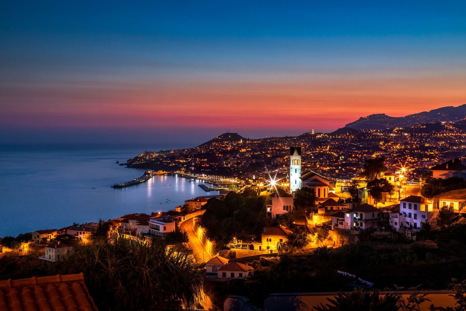 Aerial panoramic view of a coastal city illuminated at twilight with a vibrant orange and blue sunset sky.