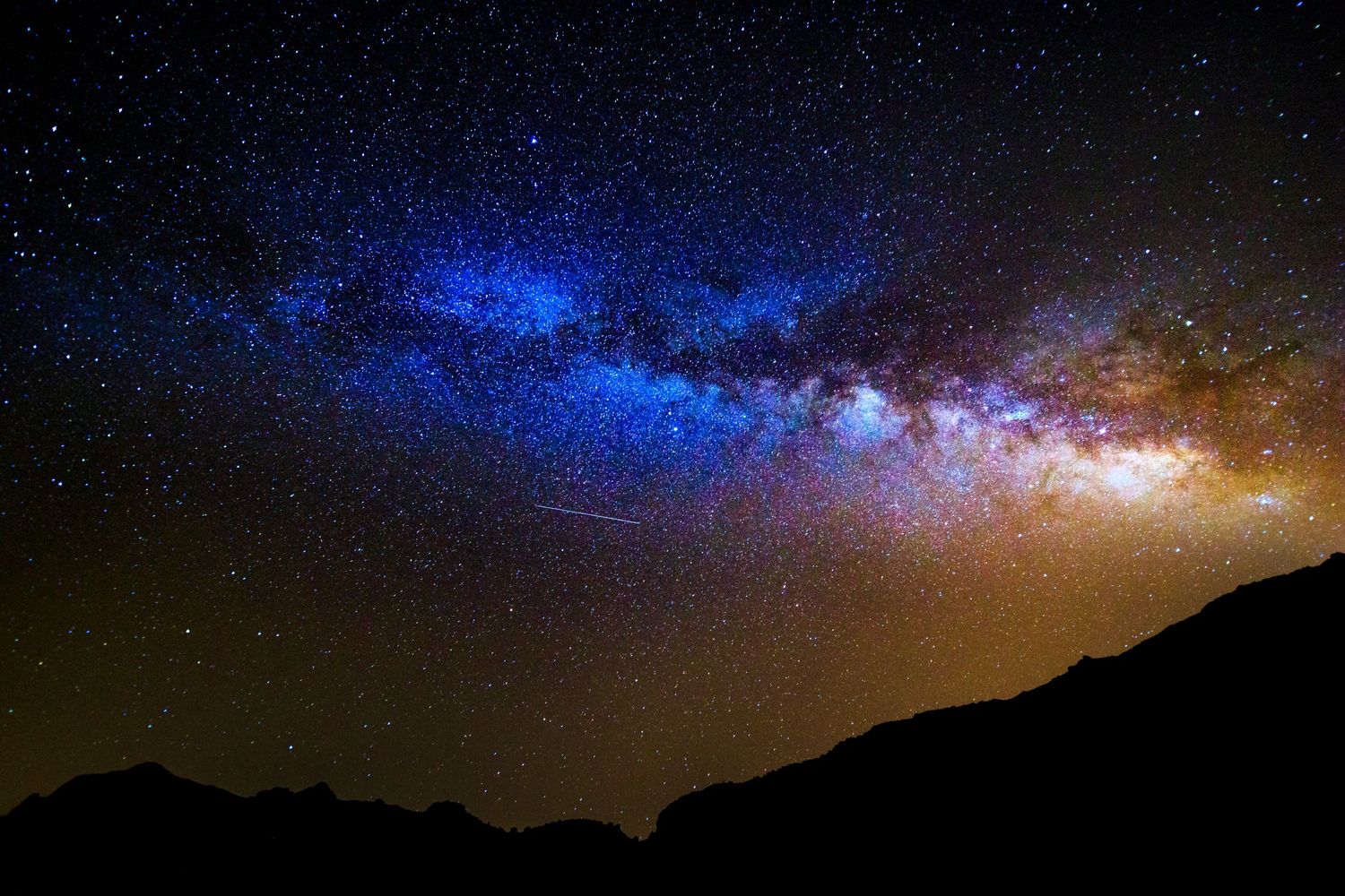 Stunning Milky Way galaxy arc over dark mountain silhouettes in a vibrant blue and gold night sky.