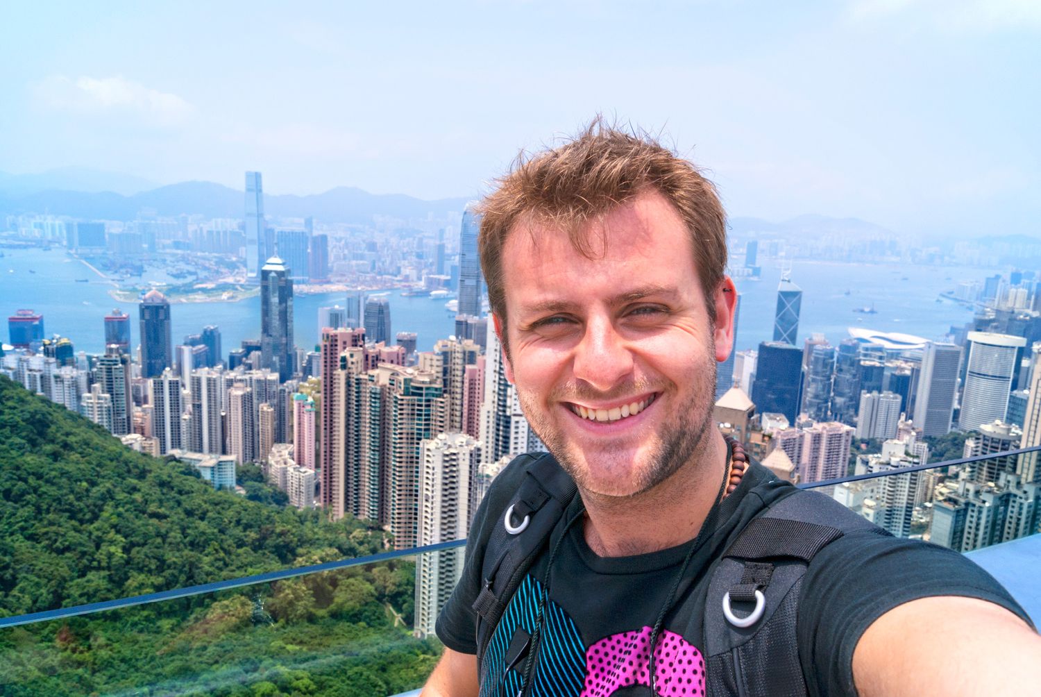 Tourist taking selfie with panoramic view of Hong Kong skyline and Victoria Harbour from Victoria Peak viewpoint.