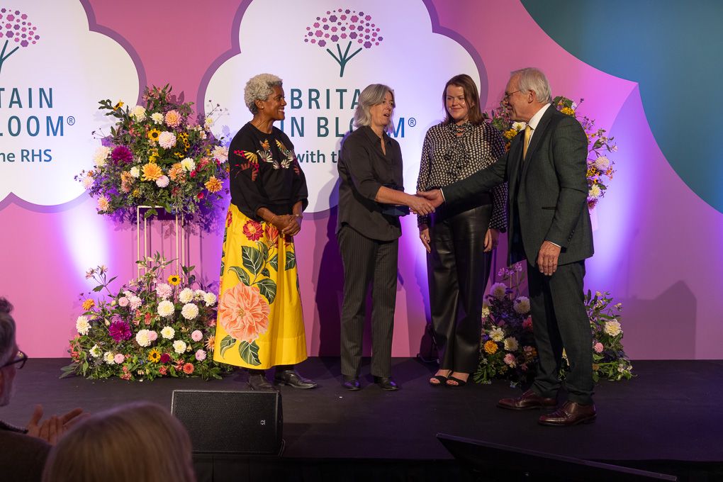 People on stage at an awards ceremony with floral decorations and pink backdrop during Britain in Bloom event.