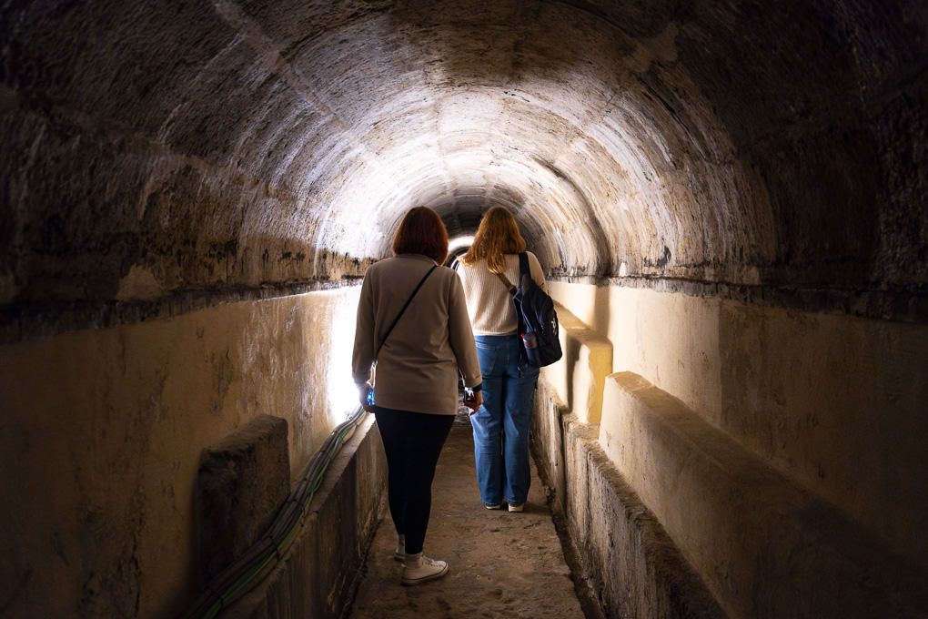 Two people walking through a dark underground tunnel with a light source visible at the end.