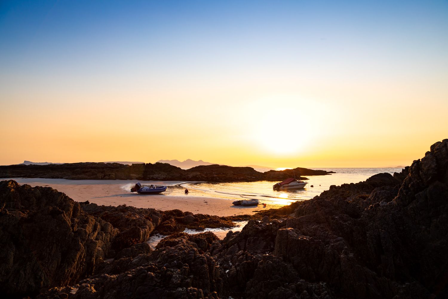 Dramatic sunset over a rocky coastal landscape with silhouetted Scottish islands and golden light reflecting on calm waters.