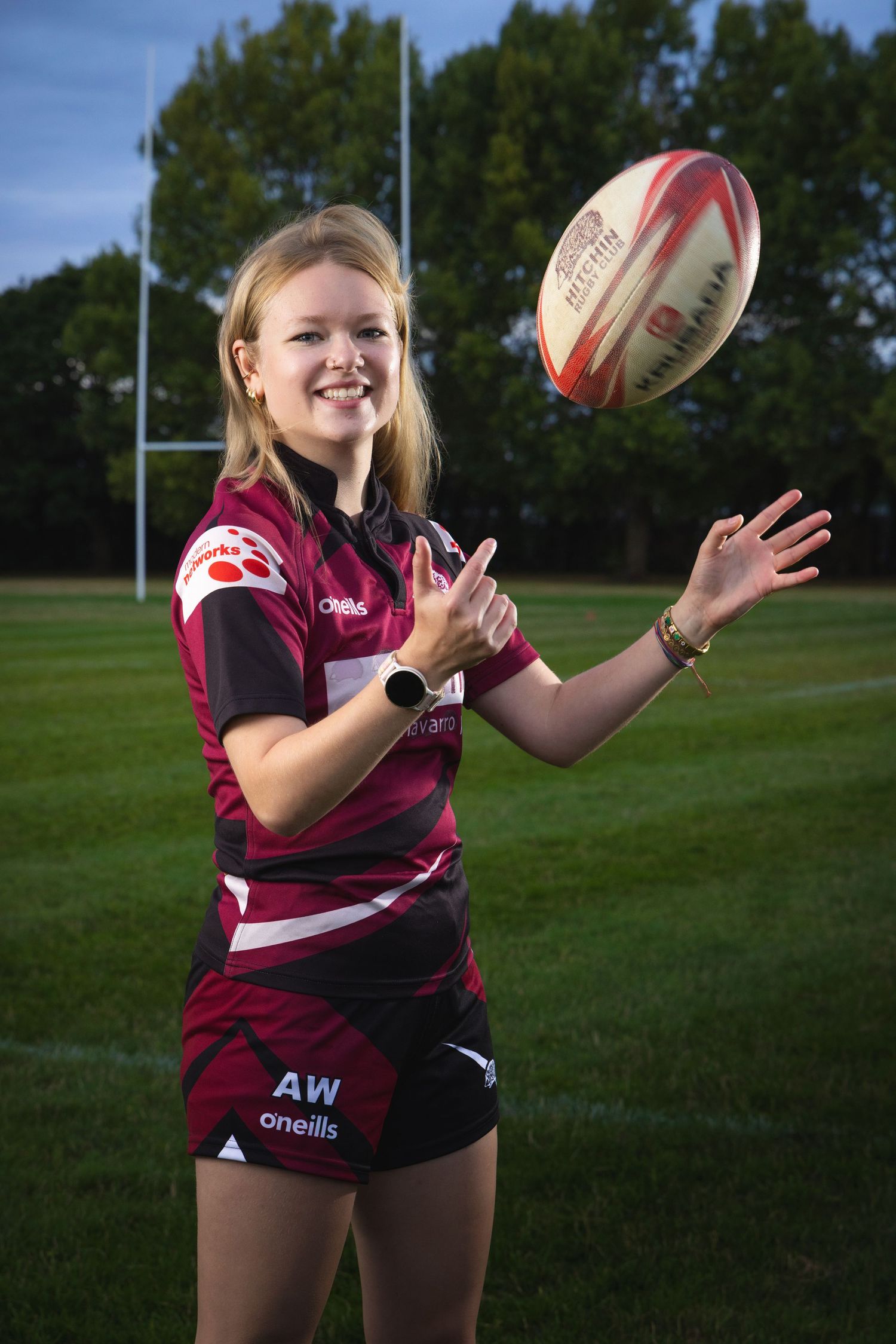Young female rugby player in maroon O'Neills kit catching a ball on a grass pitch with goalposts behind her.