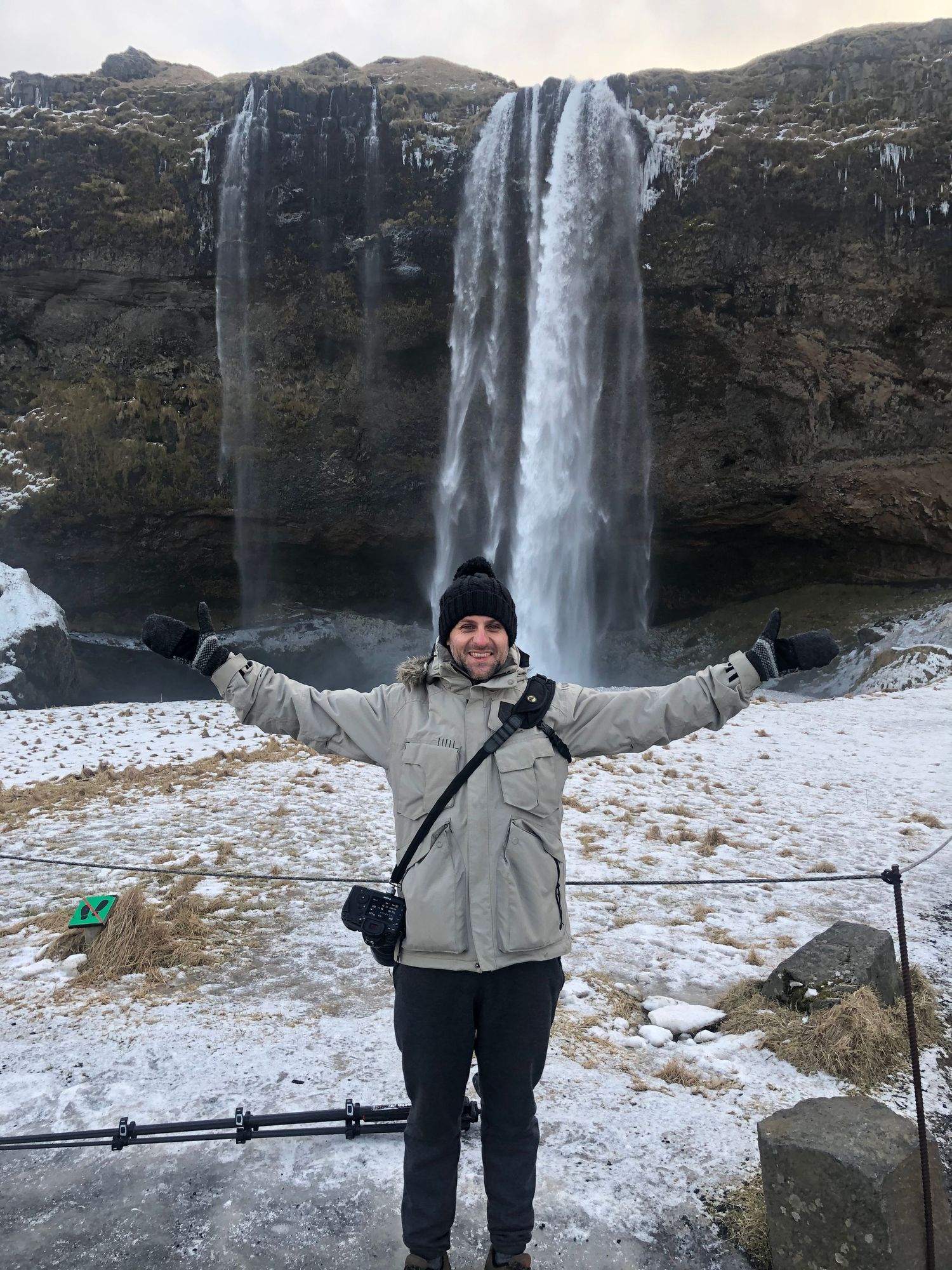 Man in winter jacket poses with arms wide open in front of a massive frozen waterfall in snowy Iceland.