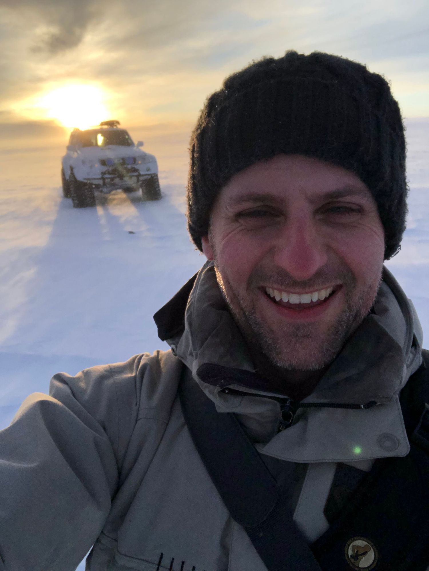 Smiling man in black winter beanie and gray jacket takes selfie on snowy landscape with SUV and sunset in background.