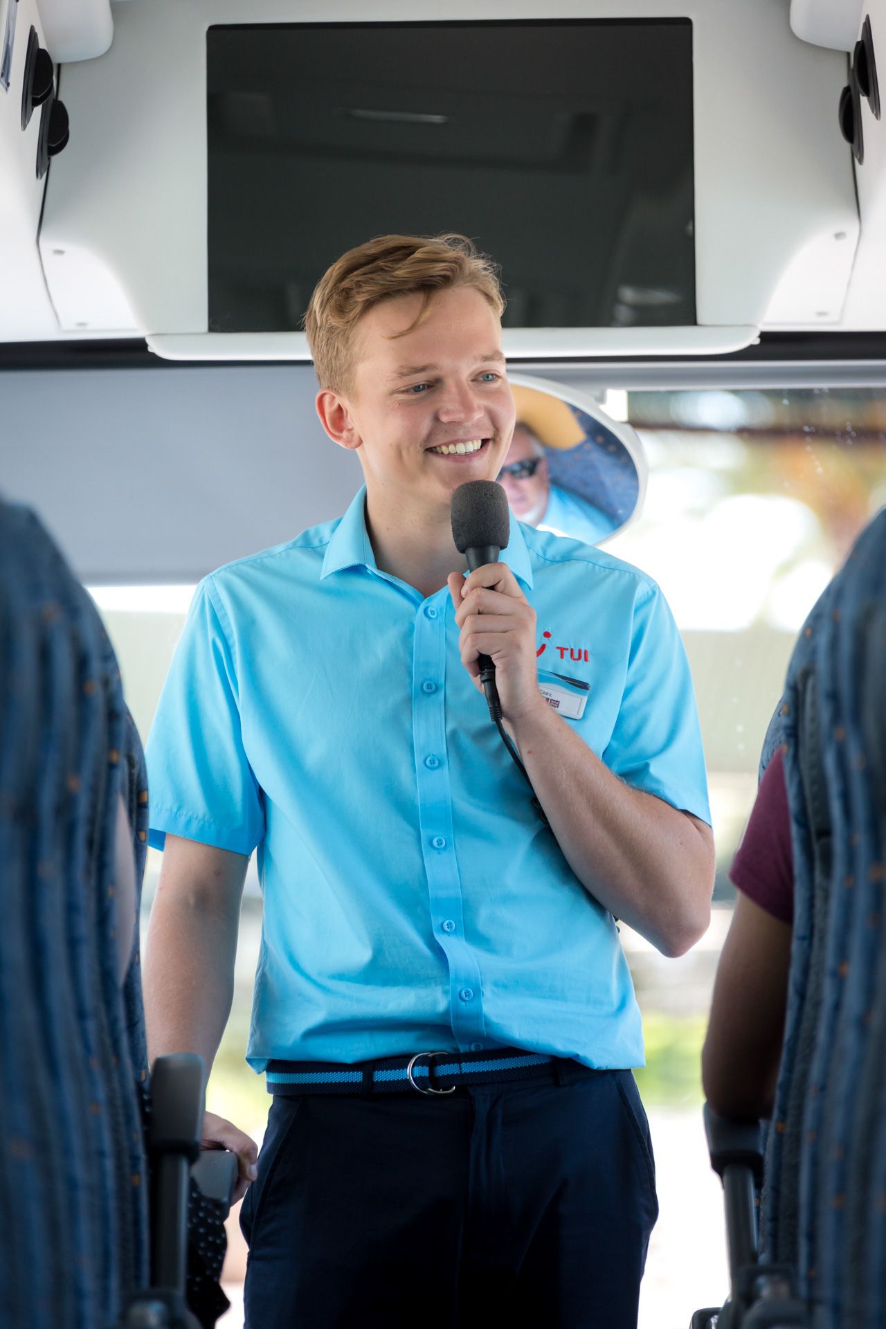 A TUI guide wearing a light blue polo shirt speaks into a microphone while addressing passengers on a bus.
