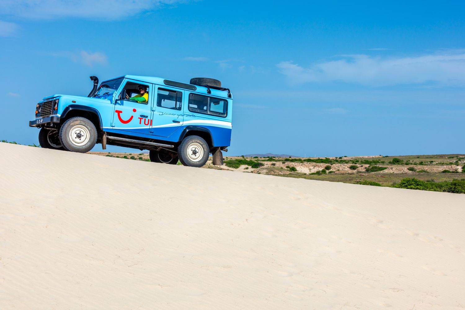 Light blue Land Rover Defender driving on a sandy dune against bright blue sky on a sunny day.